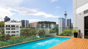 A rooftop swimming pool with a wooden deck, surrounded by glass railings, and a city skyline in the background. - Honeycomb (Auckland)