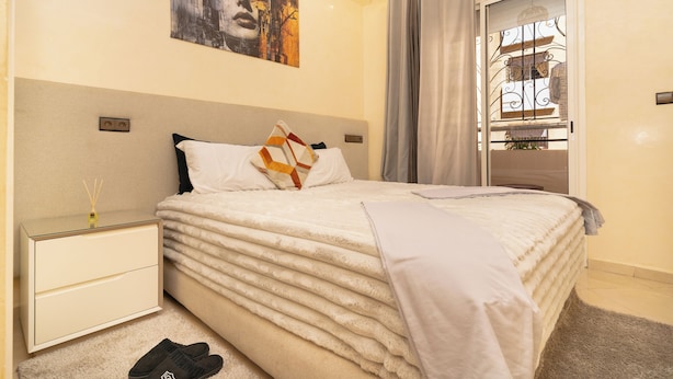 Een net opgemaakt bed met een zachte witte dekbed, een nachtkastje met een lamp en een schilderij aan de muur.