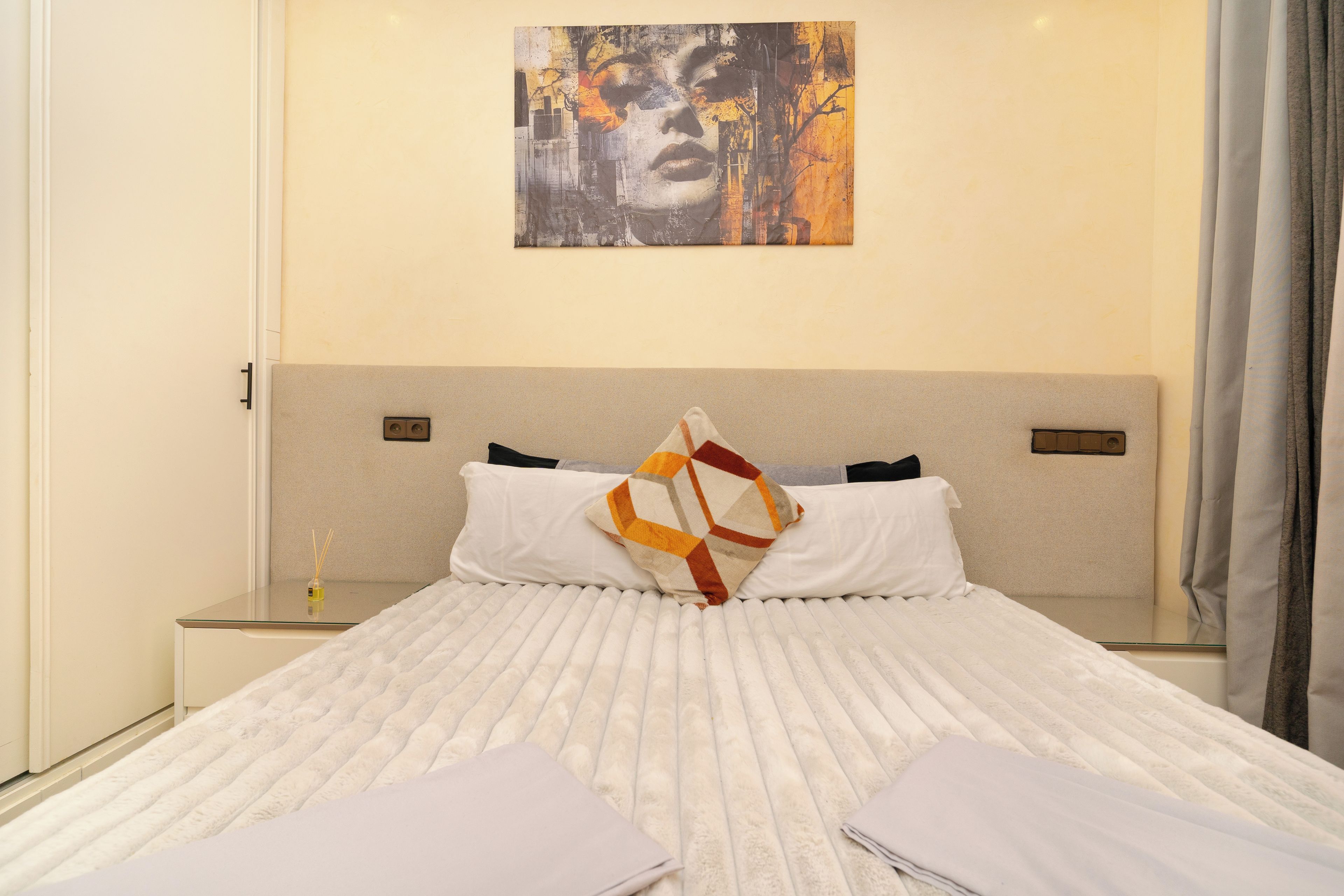 Een net opgemaakt bed met een witte deken, twee kussens en een sierkussen. Er staat een klein tafeltje met een fles en een kaars, en een schilderij aan de muur.