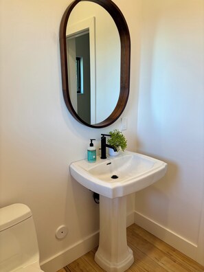 A bathroom with a white pedestal sink, a round mirror, a small potted plant, and a toilet. - Timber&Tonic Mountain Chalet—2 min to beach (Sagle)