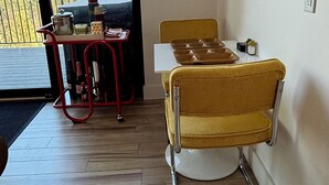 Et spiseområde med et bord og en gul stol, en rød tralle med køkkenudstyr, samt et vindue med udsigt til bjerge og træer.