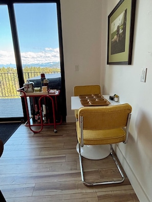 A dining area with a table and a yellow chair, a red cart with kitchen items, and a window with a view of mountains and trees. - Timber&Tonic Mountain Chalet—2 min to beach (Sagle)
