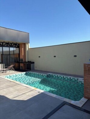Piscina moderna ao ar livre com uma fonte de água azul cristalina, rodeada por um terraço com uma espreguiçadeira e uma mesa pequena.