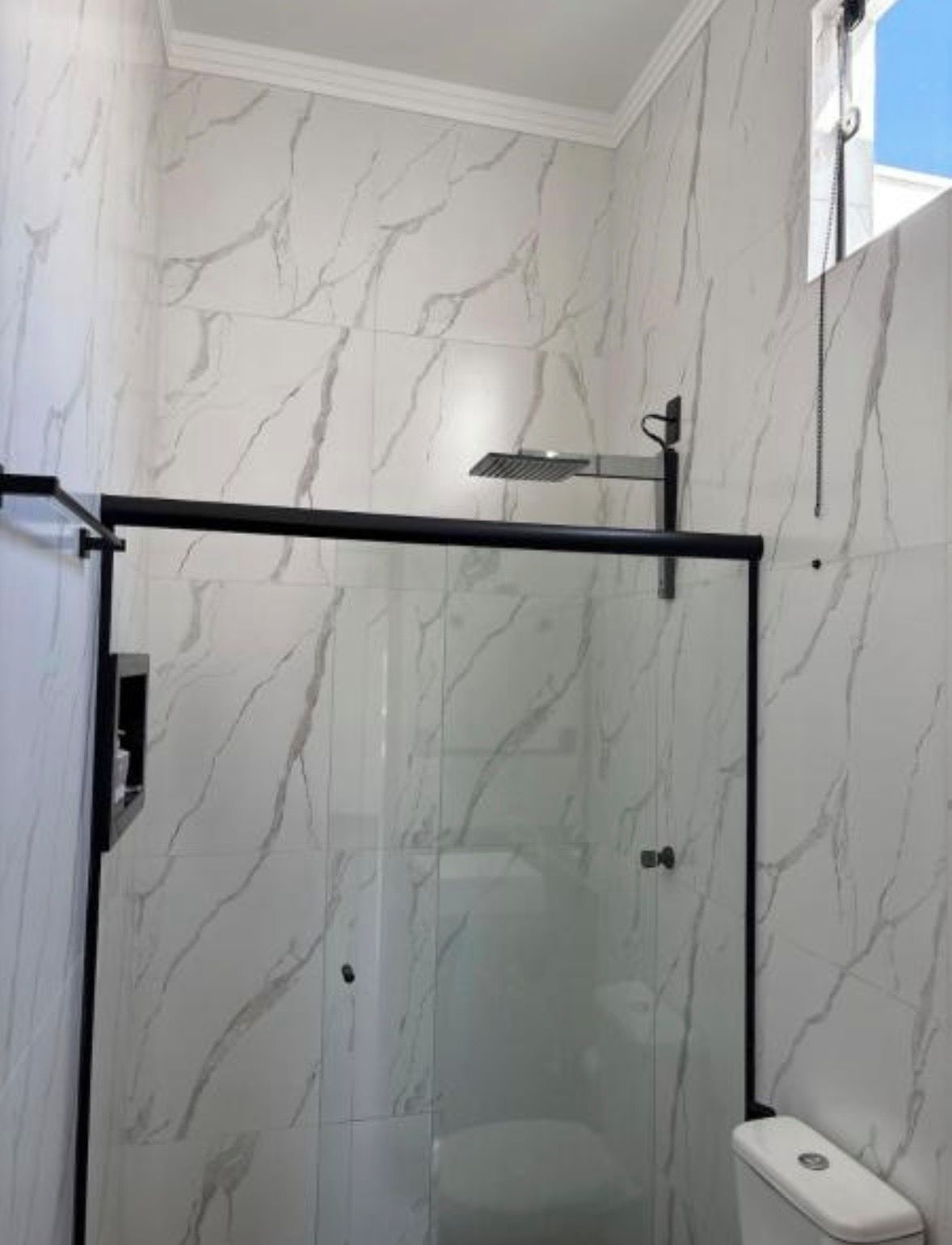 A glass-enclosed shower stall with a black metal frame, white marble walls, and a visible toilet.