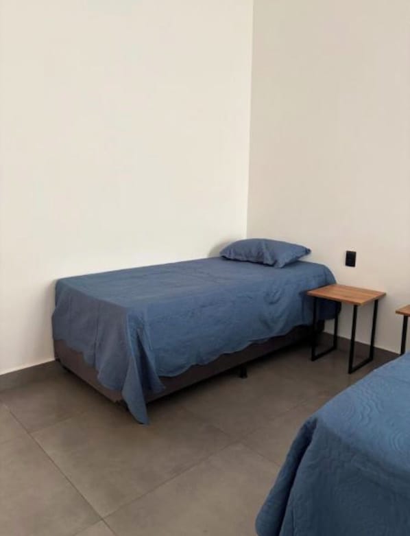 A single bed with a blue bedspread and a matching pillow in a room with plain white walls and a tiled floor.