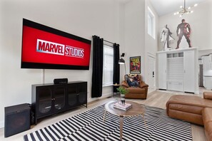 A modern living room with a large flat-screen TV displaying 'MARVEL STUDIOS,' a black entertainment unit, a brown leather sofa, a black and white striped rug, and a white cabinet with a poster. - Marvel-ous 4 BD/2.5 BA Home in Royal Oak (Royal Oak)