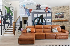 Un salon avec un canapé en cuir marron, un escalier et une fresque murale représentant des super-héros.