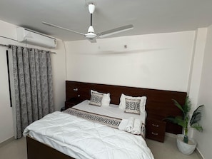 Habitación de hotel con cama, ventilador de techo, planta en maceta y ventana con cortinas.