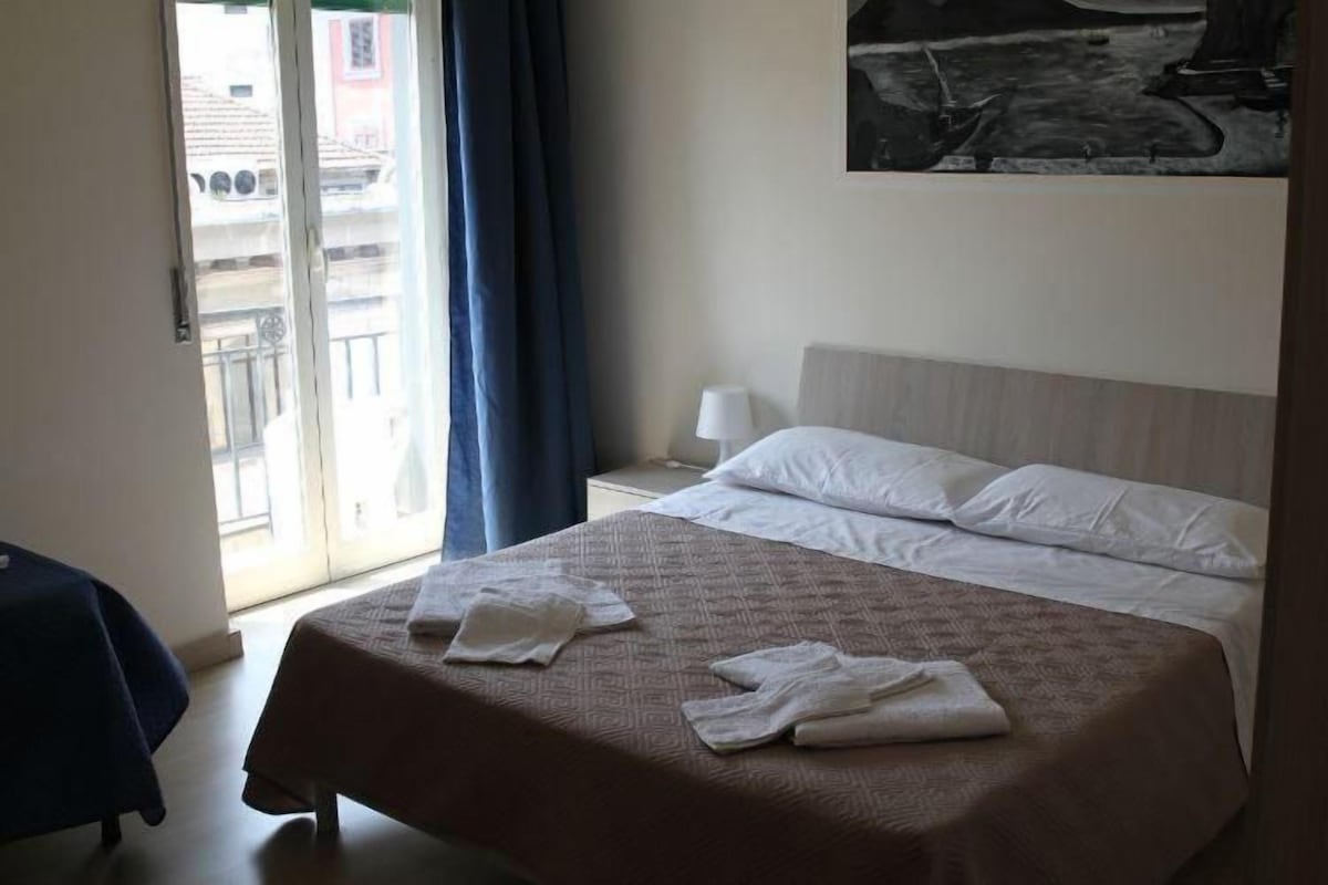 Une chambre d’hôtel avec un lit, une fenêtre avec des rideaux, une table de chevet avec une lampe et un tableau au mur.