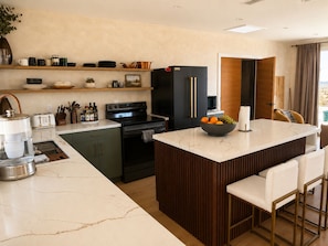 A modern kitchen with a marble countertop, a central island with bar stools, a black refrigerator, and a wooden cabinet. - Milky Way Retreat - Desert Views, Barrel Sauna , Hammock Deck & Hot Tub (Joshua Tree)