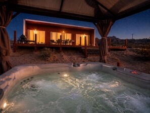 A hot tub with water, a wooden deck, and a building with lights. - Milky Way Retreat - Desert Views, Barrel Sauna , Hammock Deck & Hot Tub (Joshua Tree)