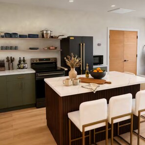 A modern kitchen with a central island, bar stools, a microwave, and a wooden door. - Milky Way Retreat - Desert Views, Barrel Sauna , Hammock Deck & Hot Tub (Joshua Tree)