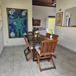A dining area with a wooden table and chairs, a large abstract painting on the wall, and a view of the outdoors through a door. - Charming beachfront villa, right on the water's edge (La Trinité)