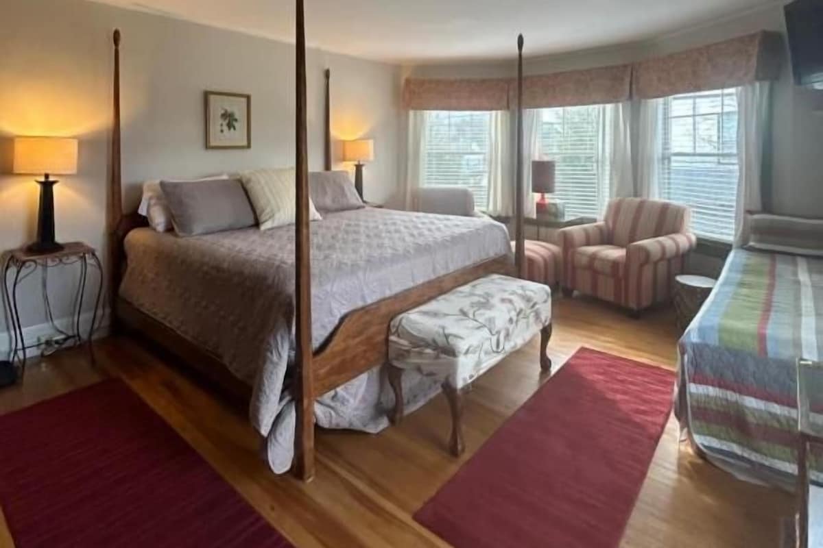 Quarto com cama de dossel, um banco, uma cadeira e uma mesinha.