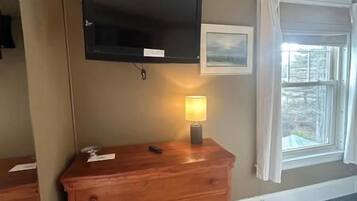 A room with a wooden dresser, a flat-screen TV mounted on the wall, a bedside lamp, and a window with a view.