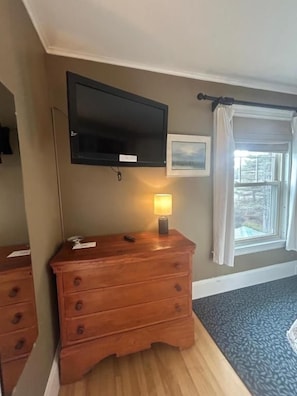 Una habitación con tocador de madera, televisor de pantalla plana montado en la pared, lámpara de noche y ventana con vistas.
