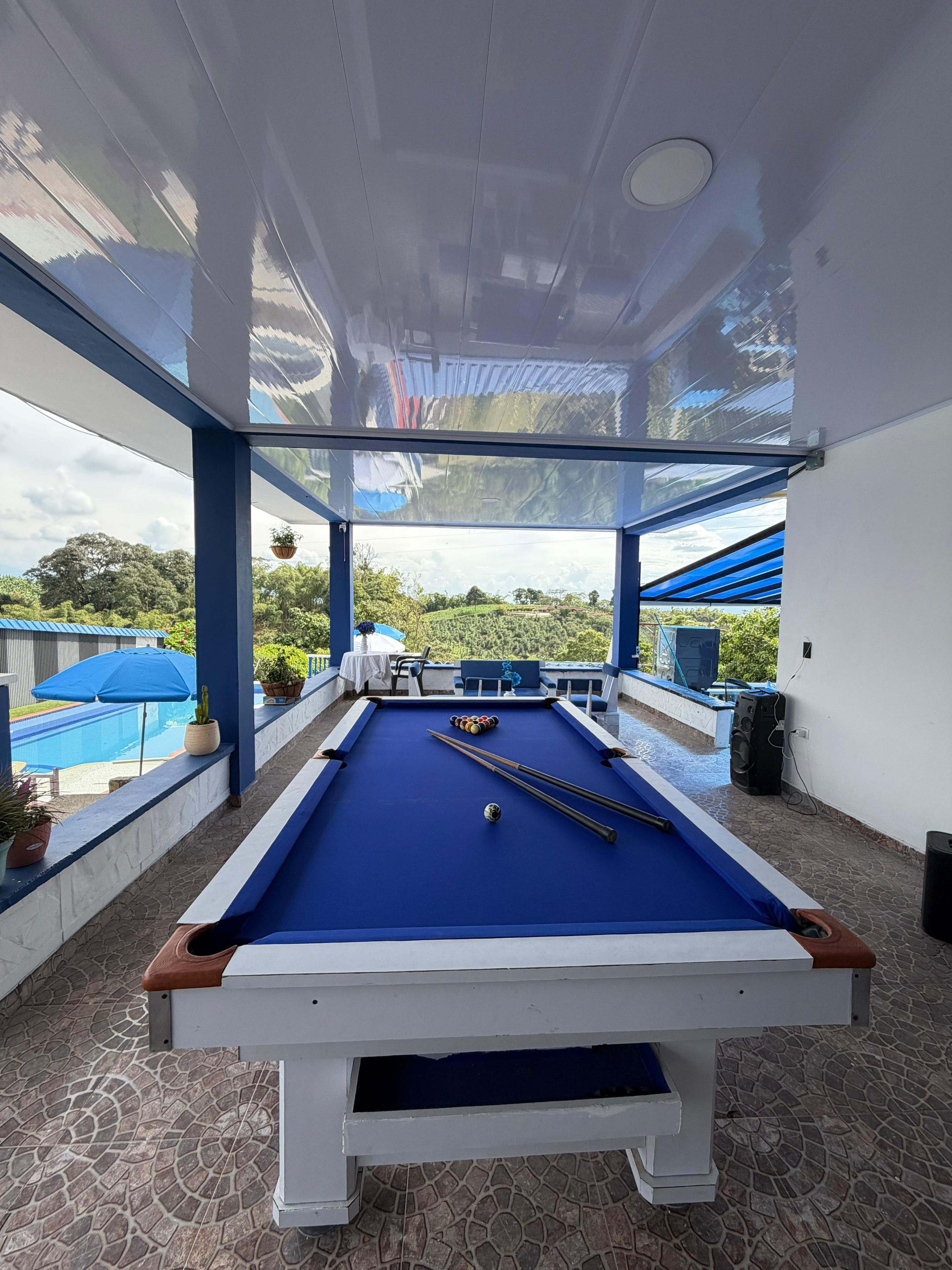 Una mesa de billar con superficie de fieltro azul, rodeada por un suelo de baldosas de piedra, y vistas a una piscina y vegetación al exterior.