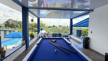 Una mesa de billar con superficie de fieltro azul, rodeada por un suelo de baldosas de piedra, y vistas a una piscina y vegetación al exterior.