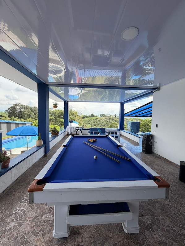 A pool table with a blue felt surface, surrounded by a stone-tiled floor, and a view of a pool and greenery outside.