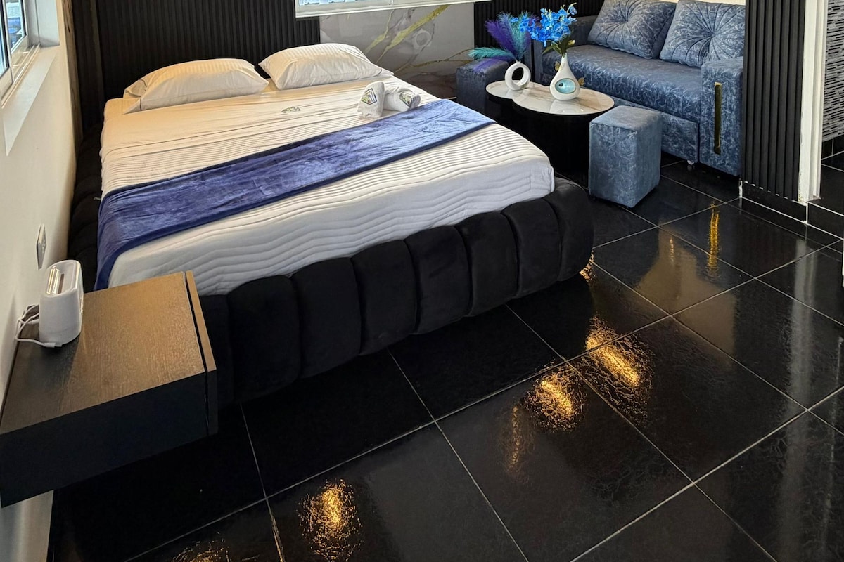 A modern bedroom with a large bed, a bedside table, a small round table with a vase, and a view of greenery outside.