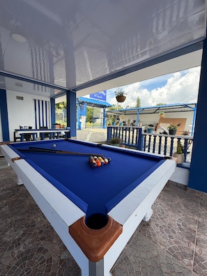 An indoor pool table with a blue felt surface, surrounded by a blue and white color scheme, and a view of the outside through an open door. - Artehotel del Quindío  (Filandia)