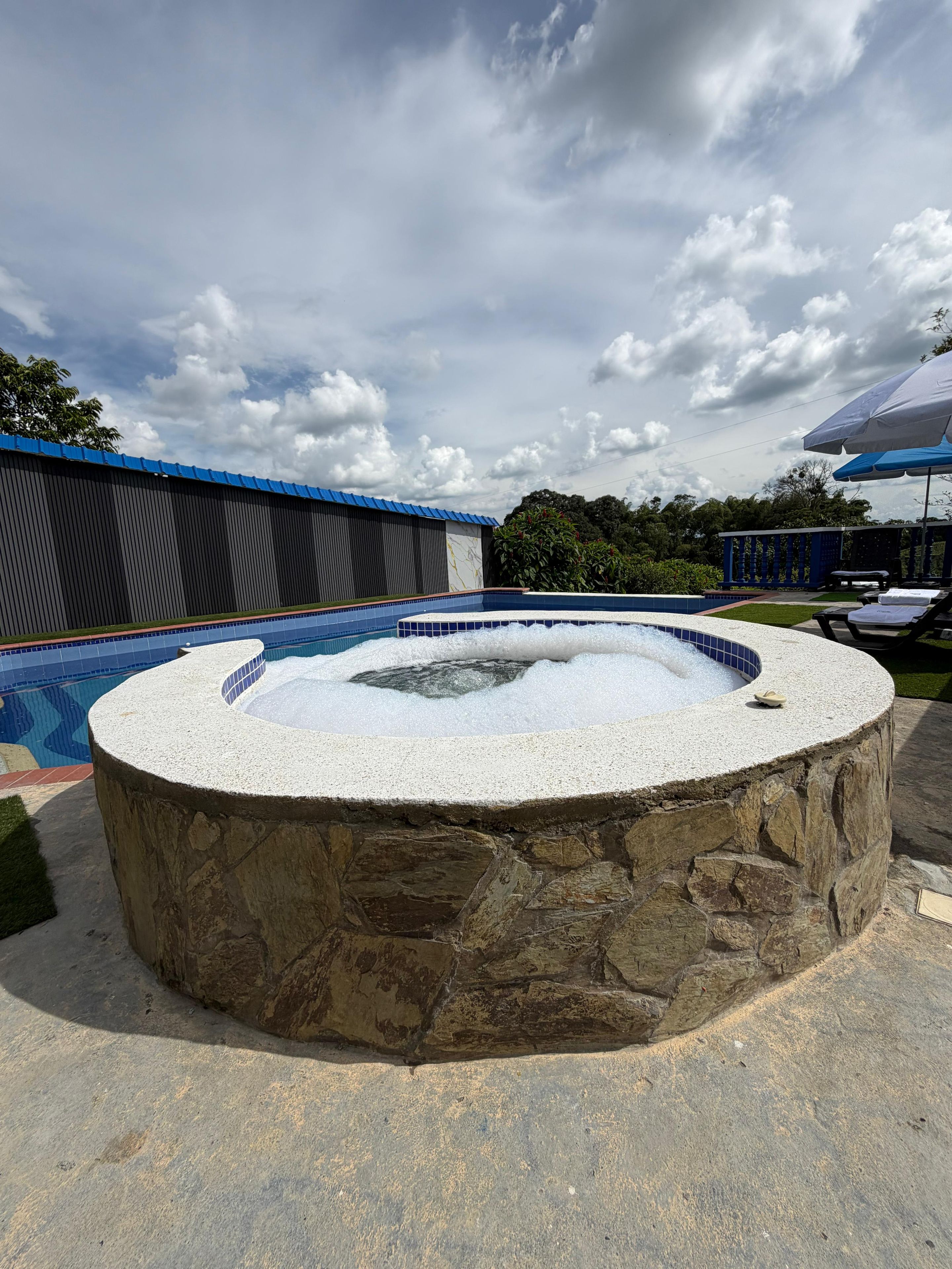 Un jacuzzi rodeado de muros de piedra, con una piscina y zona de descanso al fondo.