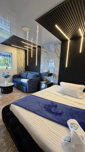 A modern bedroom with a large bed, a sofa, a small table with a vase, and a window with a view of greenery.