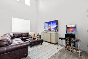 Ein Wohnzimmer mit braunem Ledersofa, einem wandmontierten Flachbildfernseher, einer Kommode und einer Spielhalle.