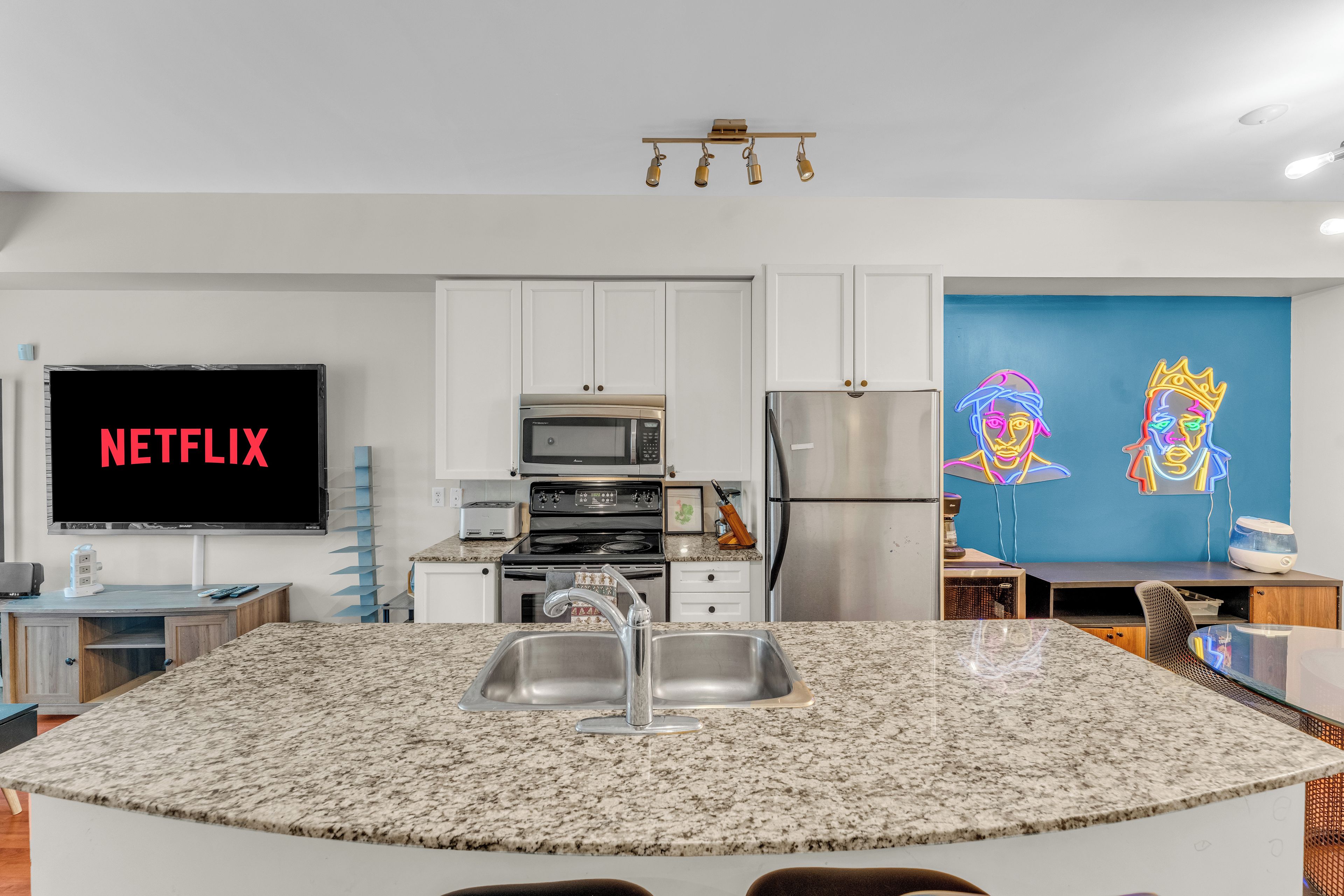 A modern kitchen with a granite countertop, stainless steel appliances, and a flat-screen TV displaying 'NETFLIX'.