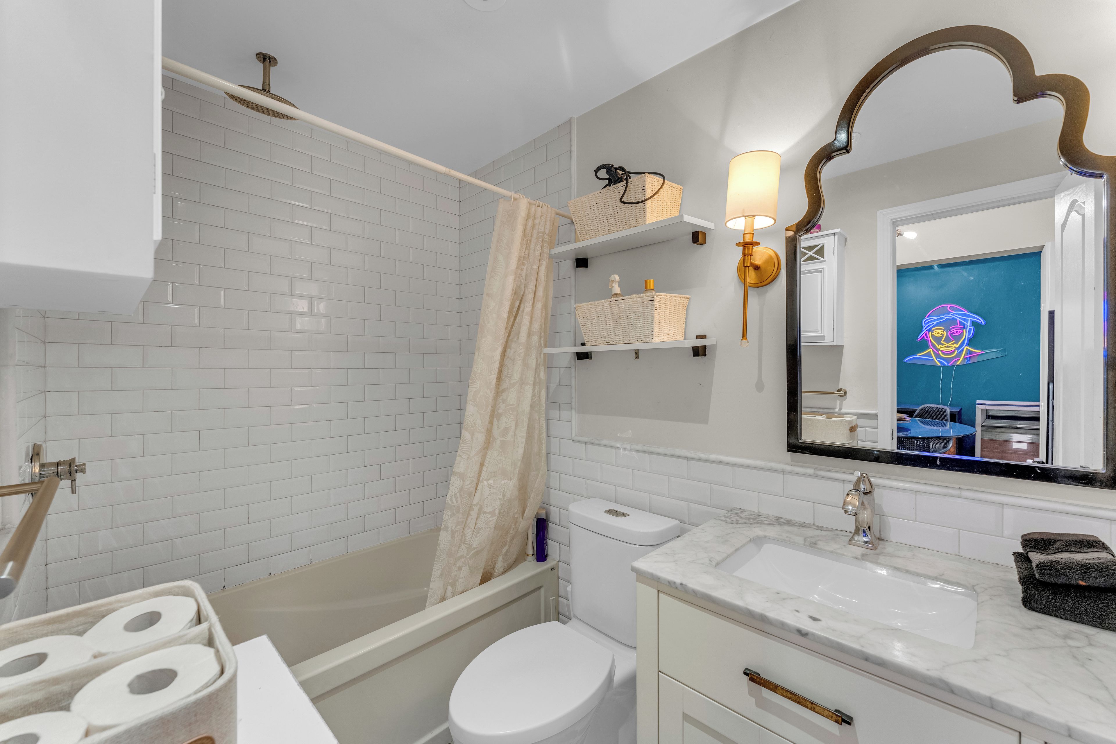 Un baño con paredes de azulejos blancos, una bañera, un inodoro, un lavabo con encimera de mármol y un espejo con marco decorativo.