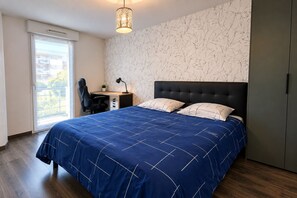 A bedroom with a large bed, a desk, and a chair. There is a window with a view of greenery outside.