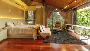 Ein Schlafzimmer mit einem Bett, einem Sessel, einem Schreibtisch und einem großen Fenster mit Blick auf Grünflächen.