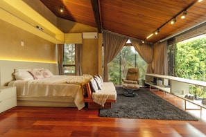 Een slaapkamer met een bed, een stoel, een bureau en een groot raam met uitzicht op groen.