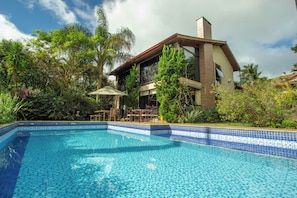 Een zwembad met betegelde rand, omgeven door weelderige begroeiing en een gebouw met twee verdiepingen en een balkon.