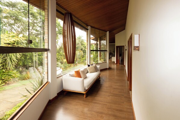 A modern interior with a wooden floor, a white sofa with cushions, and large windows offering a view of greenery.