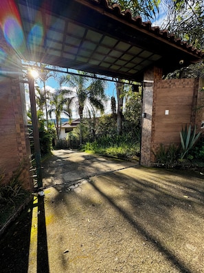 A covered entrance to a property with a paved driveway leading to a building, surrounded by greenery and palm trees.
