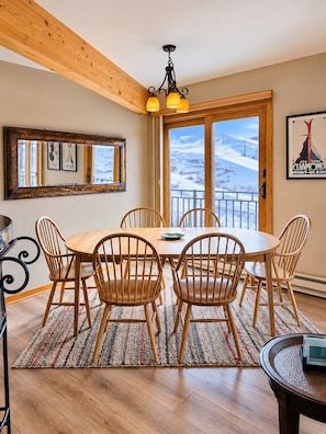 A dining area with a wooden table and chairs, a chandelier, a large mirror, and a view of a snowy landscape through a glass door. - True Ski-In/Ski-Out, Mountain Views with Garage & Elevator (Mt Crested Butte)