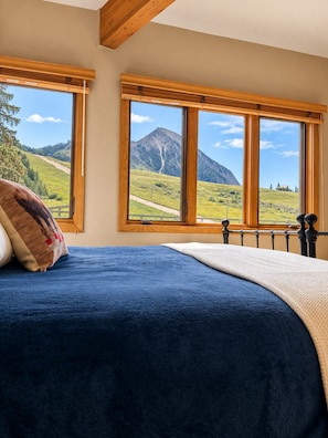 Une chambre avec un lit, deux fenêtres donnant sur les montagnes, des poutres en bois au plafond et une vue sur un paysage verdoyant.