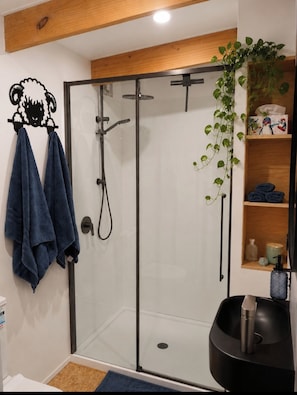 Ein modernes Badezimmer mit einer Duschkabine aus Glas, einem Handtuchhalter mit zwei Handtüchern, einer Ablage mit Pflegeprodukten und einer hängenden Pflanze.