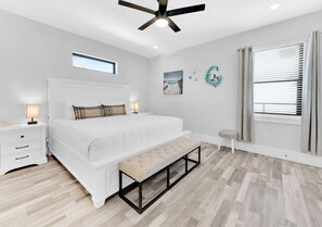 Un dormitorio con cama, mesita de noche, banqueta, ventilador de techo, obra de arte, ventana con persianas y un taburete pequeño.