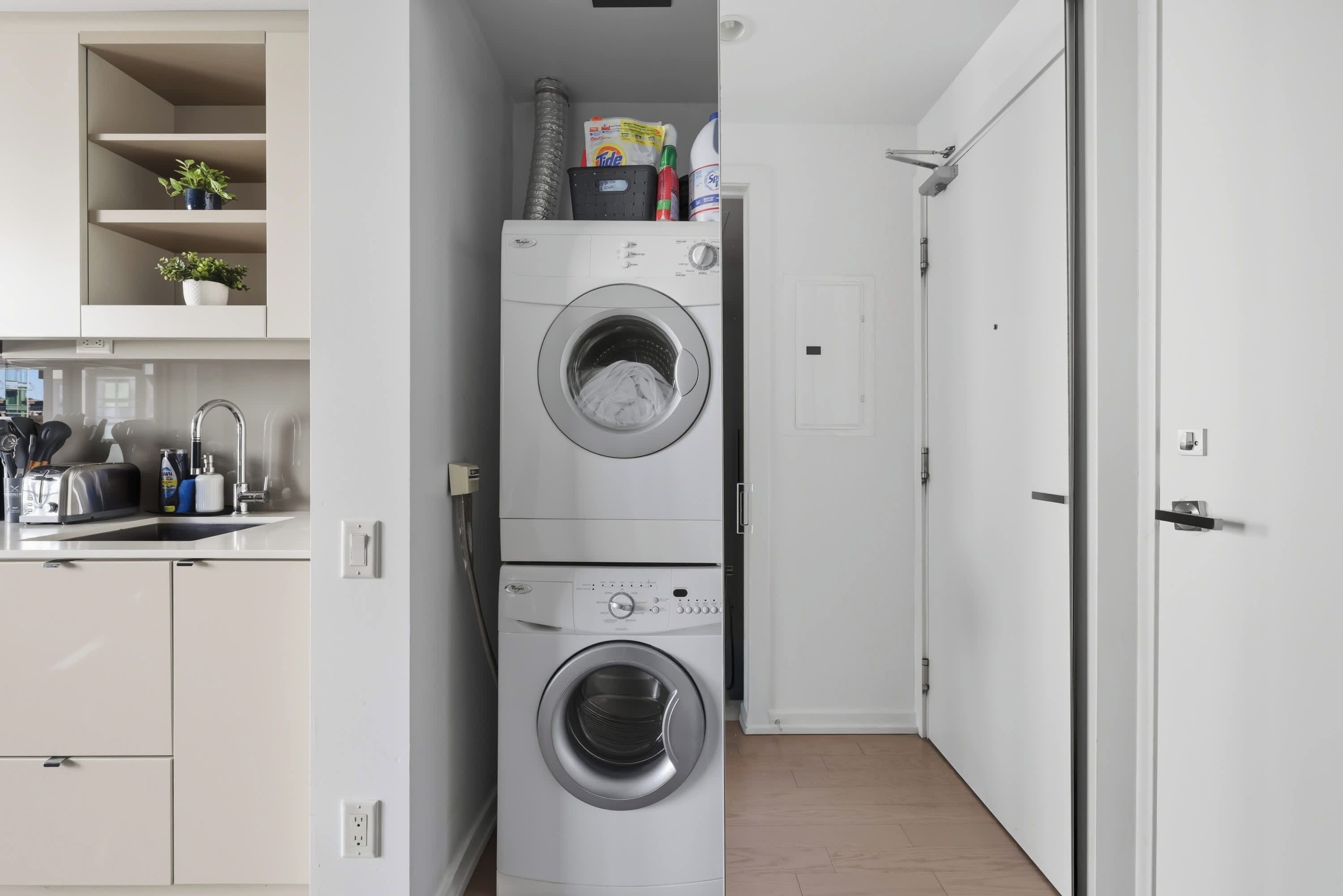 A compact laundry room with stacked washing machines, a small kitchen area with a toaster, and a door leading to another room.