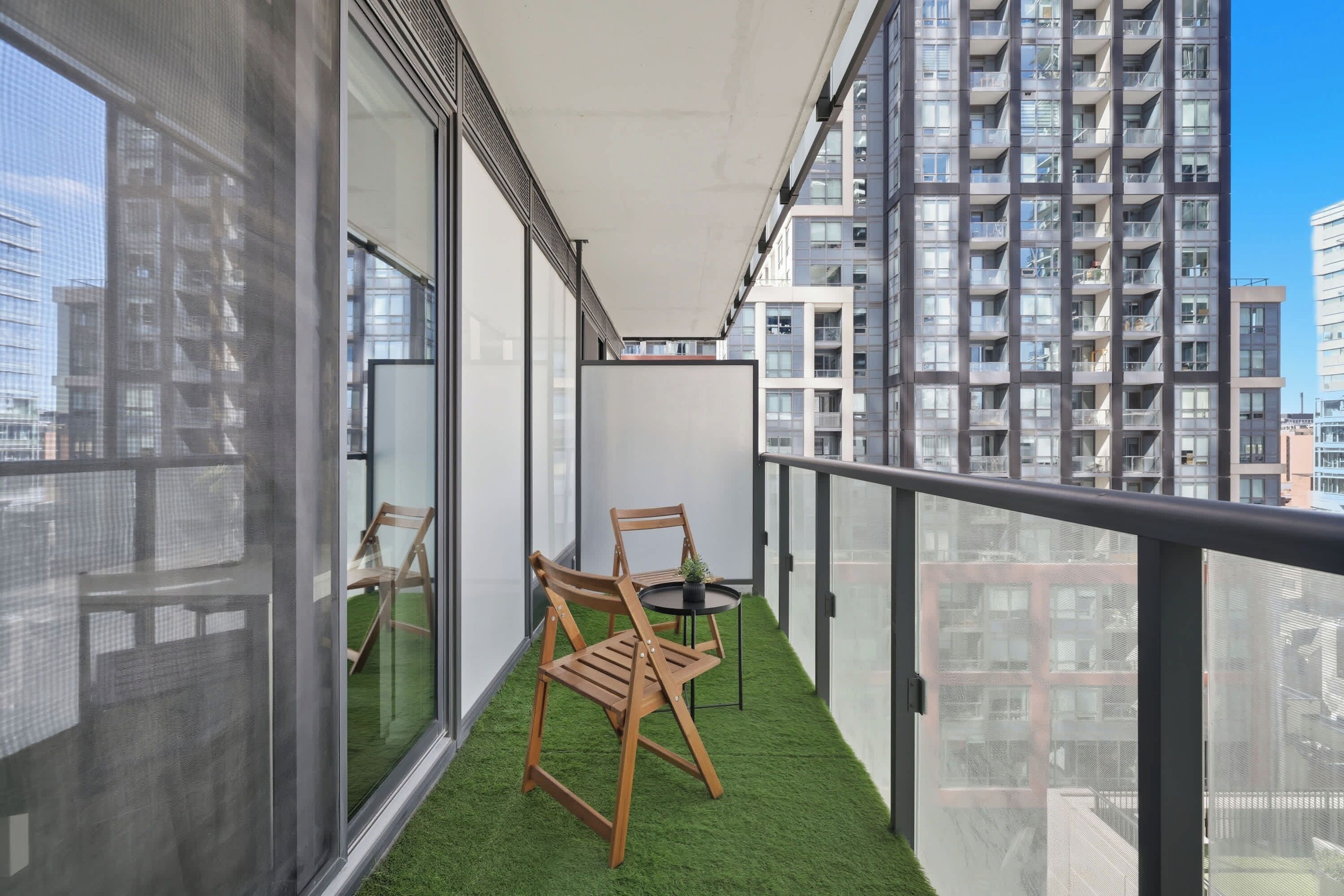 A balcony with a small table and two chairs, surrounded by glass walls and a view of high-rise buildings.