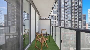 A balcony with a small table and two chairs, surrounded by glass walls and a view of high-rise buildings.