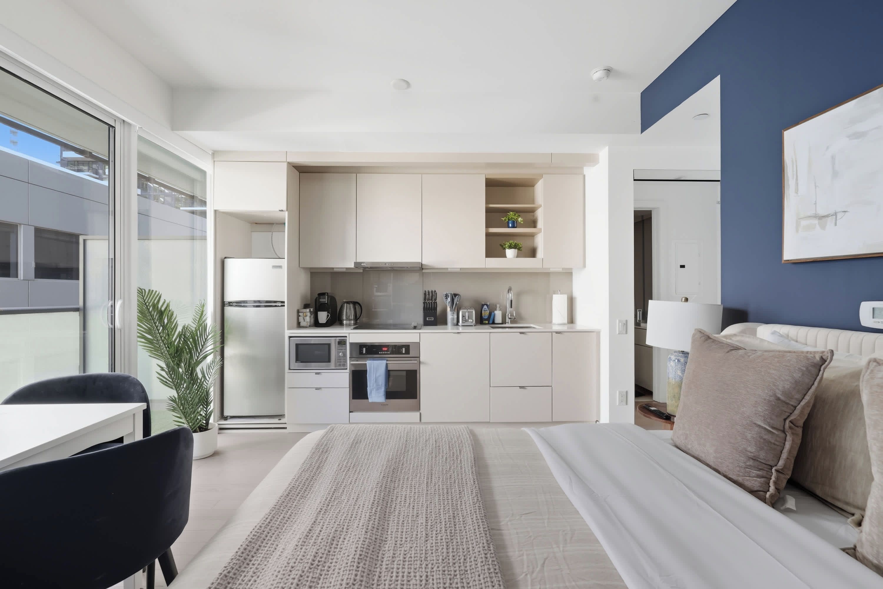 A modern hotel room with a kitchenette, a bed with a large window, and a view of the city.
