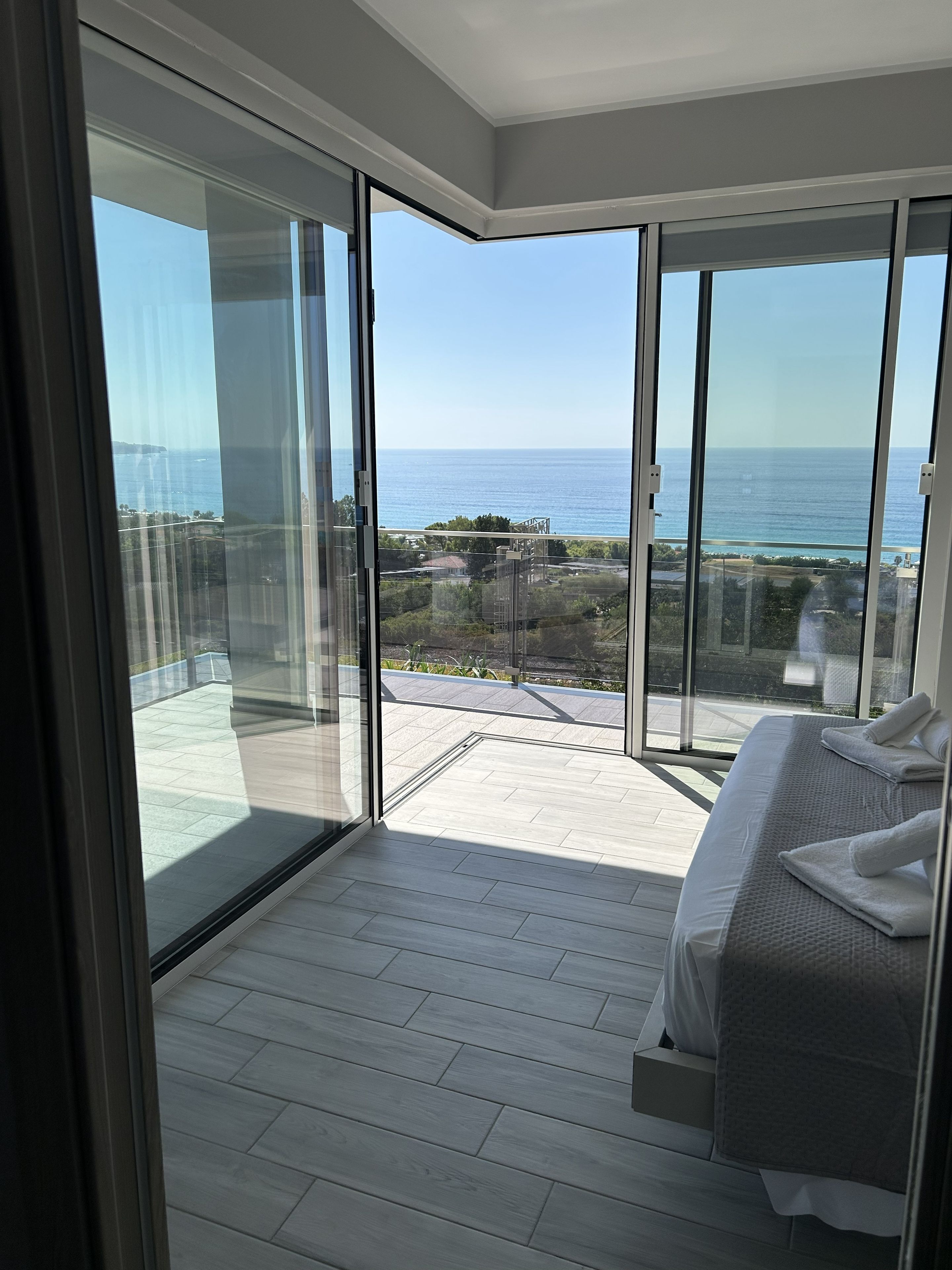 A modern bedroom with a large sliding glass door leading to a balcony with a view of the sea and distant buildings.