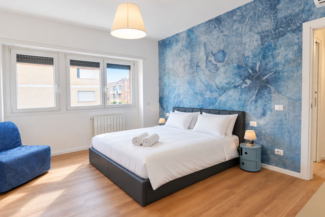 Une chambre avec un grand lit, une chaise bleue, une fresque murale et une fenêtre avec des stores.