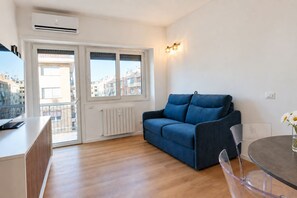 Un salon moderne avec un canapé bleu, une table à manger avec des chaises, un radiateur et une fenêtre donnant sur des immeubles.