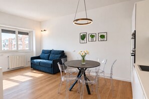 A modern living room with a blue sofa, a round dining table with clear chairs, a hanging light fixture, and framed cactus artwork on the wall. - Elegantis Trastevere Family Apartment with Terrace (Roma)