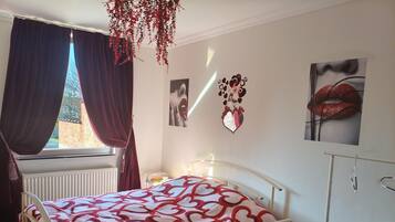 A bedroom with a bed, a radiator, a window with curtains, wall art, and a bedside table.