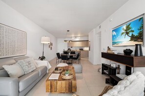 A modern living room with a sofa, coffee table, and TV. There's a kitchen in the background with a dining area. - 23rd Floor Double Condo Ocean Views and Pools (Hollywood)